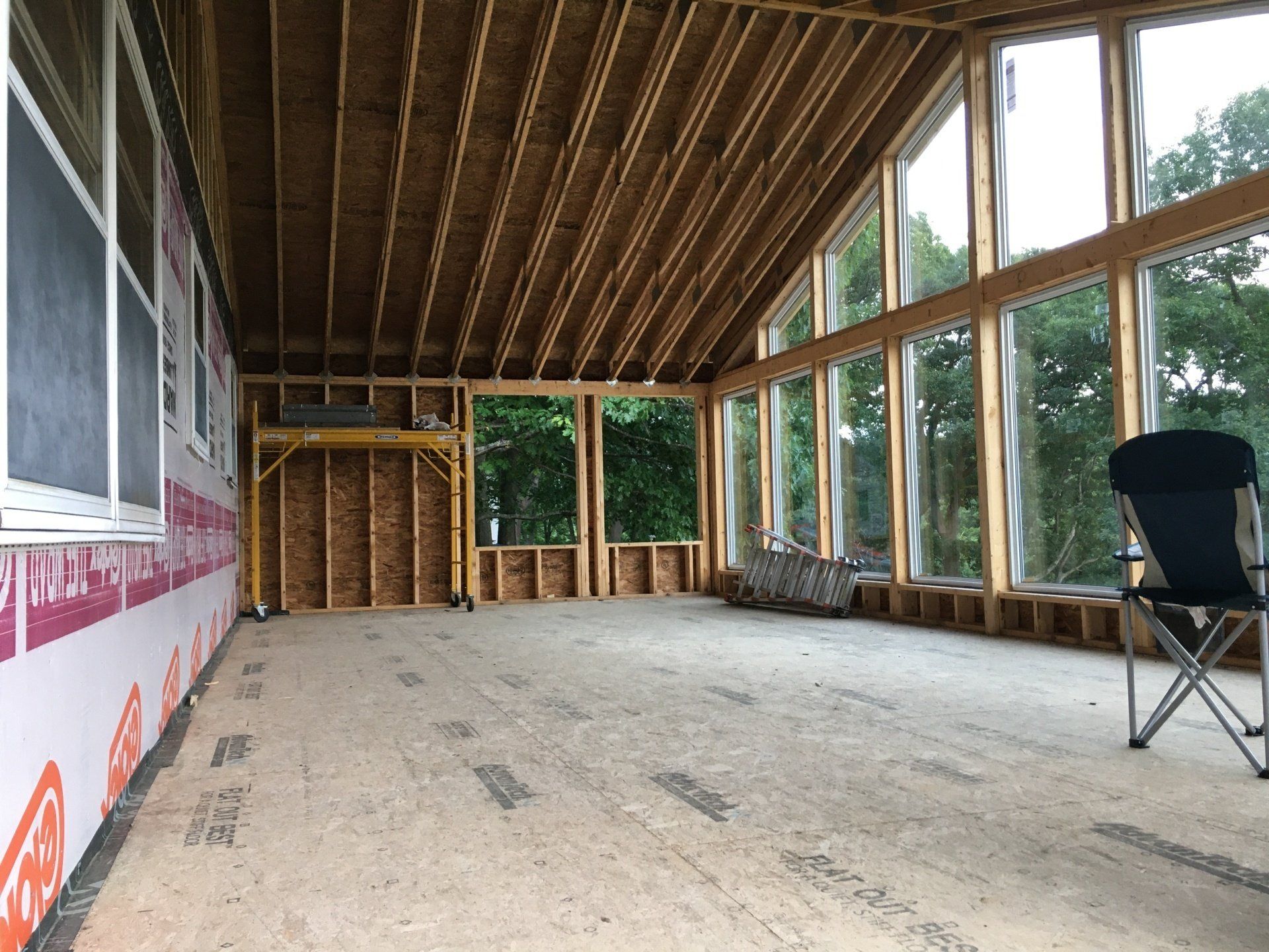Unfinished enclosed porch with exposed wood framing, large windows, and a folding chair on the right.