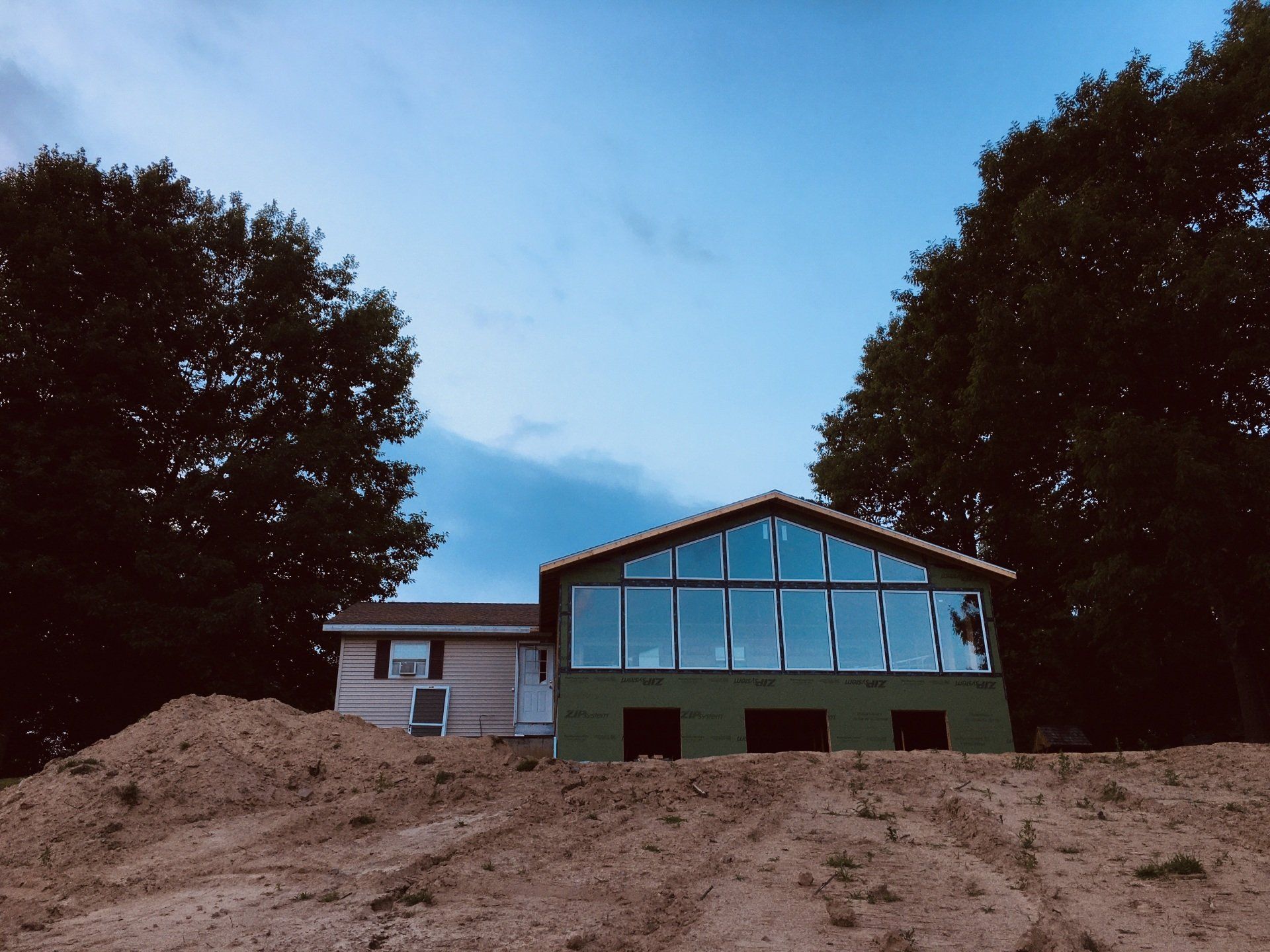 House under construction with large windows, framed by two trees and a mound of dirt in front