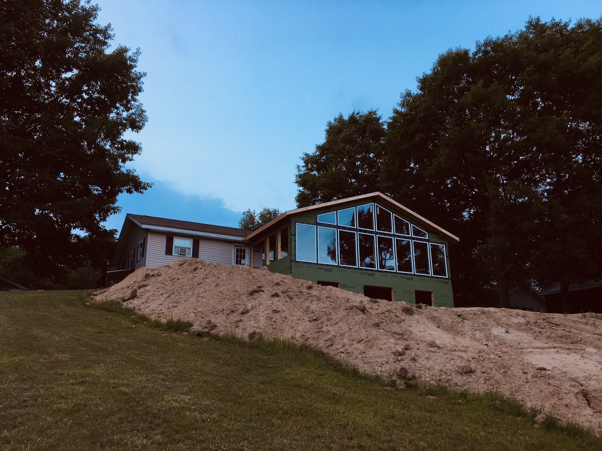 Modern house on a grassy hill with a large dirt mound, surrounded by trees under a blue sky