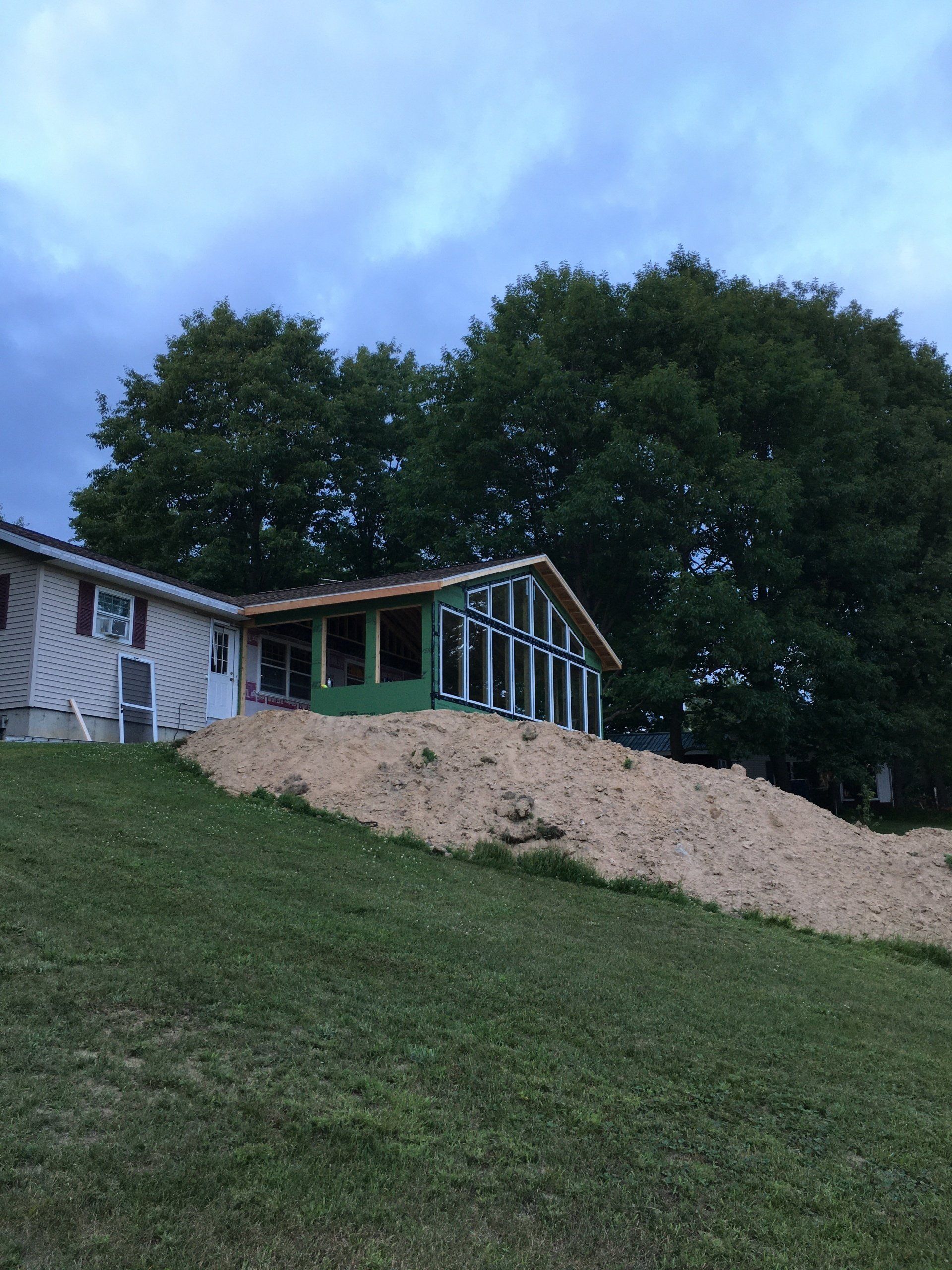 House with green porch on a grassy hillside at dusk, backed by tall trees