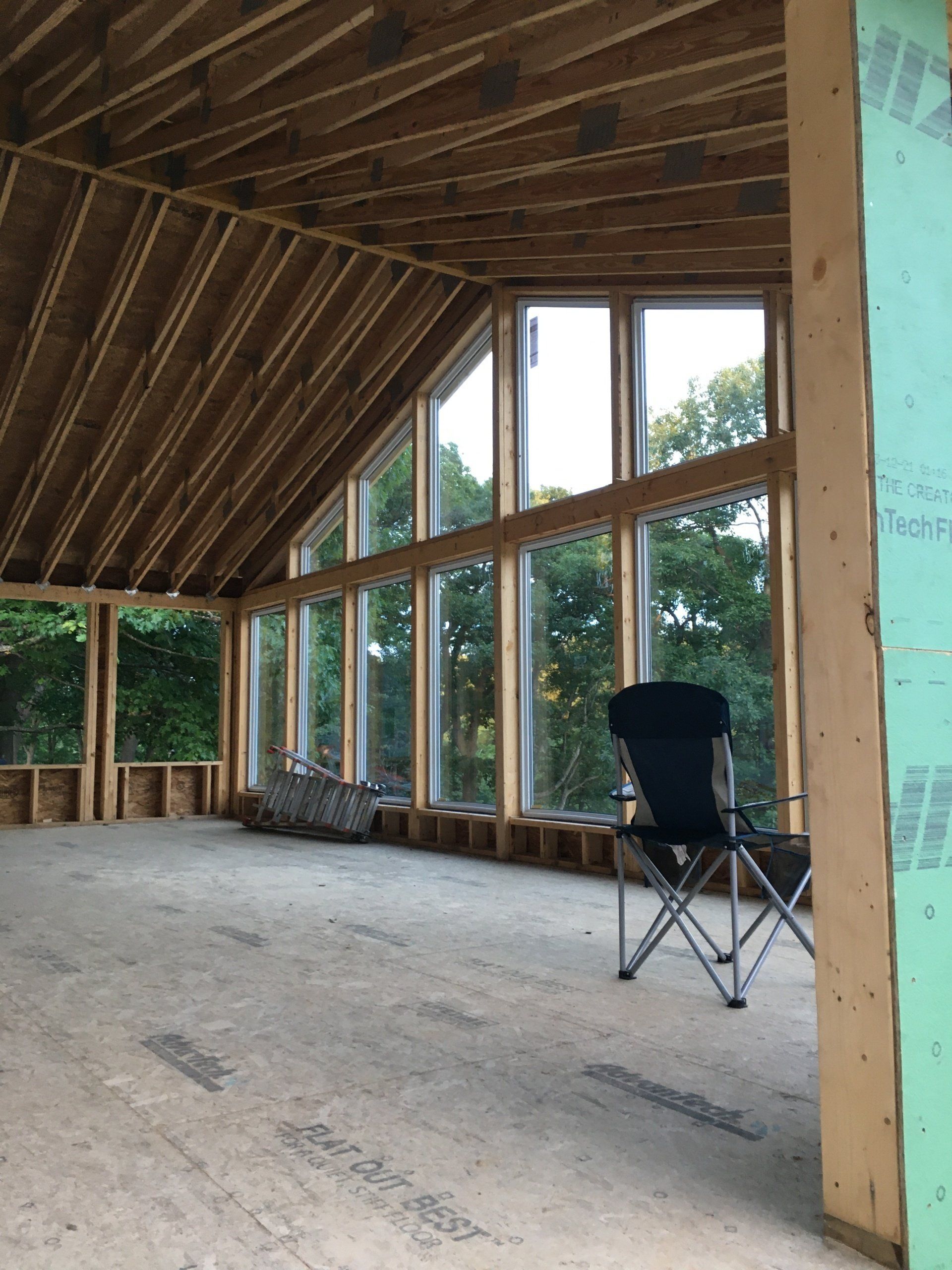 Unfinished timber-framed room with large windows, exposed roof trusses, and a folding chair on a concrete floor