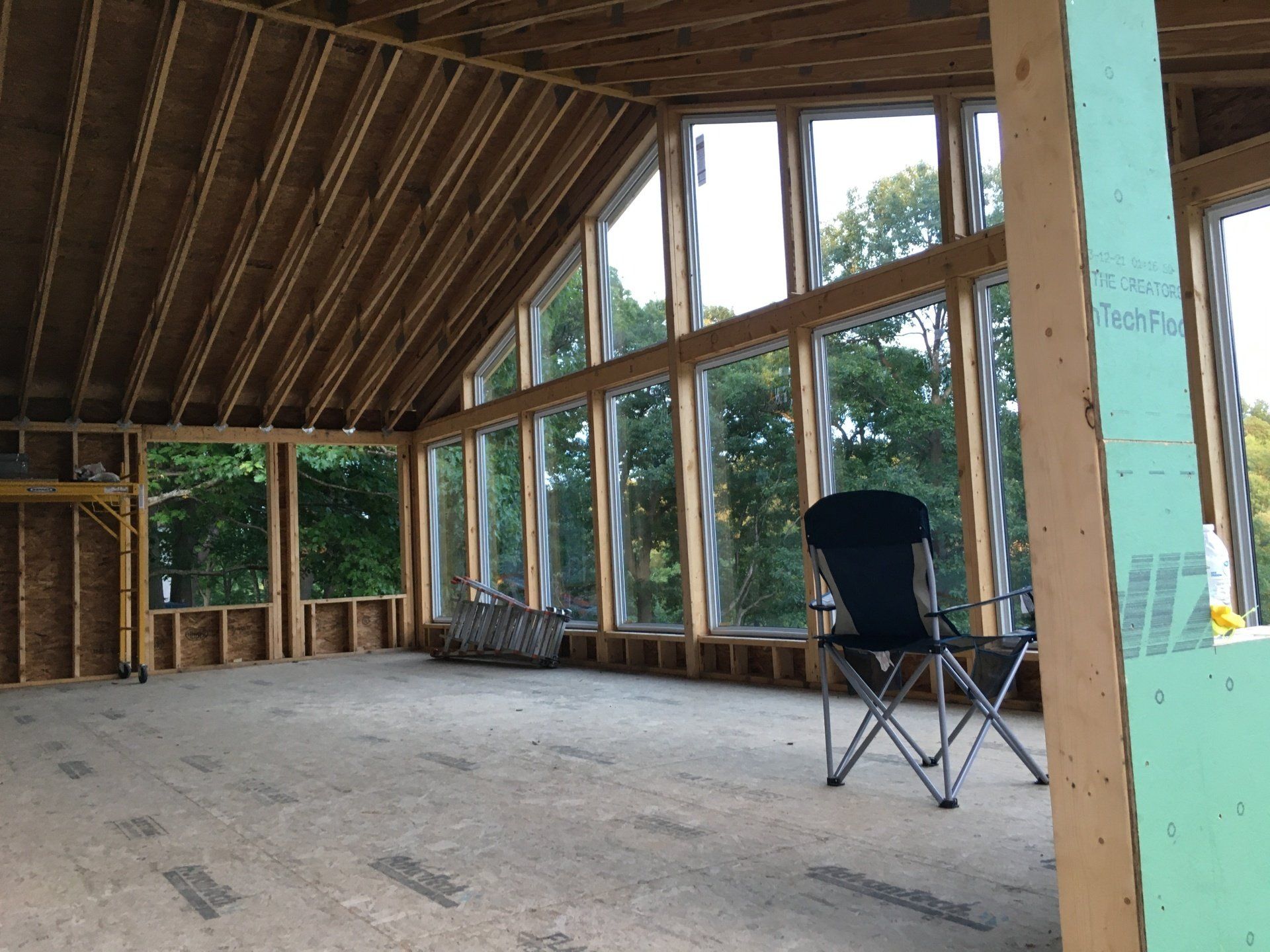 Unfinished room with exposed wooden framing, slanted ceiling, large windows, and a folding chair on the concrete floor