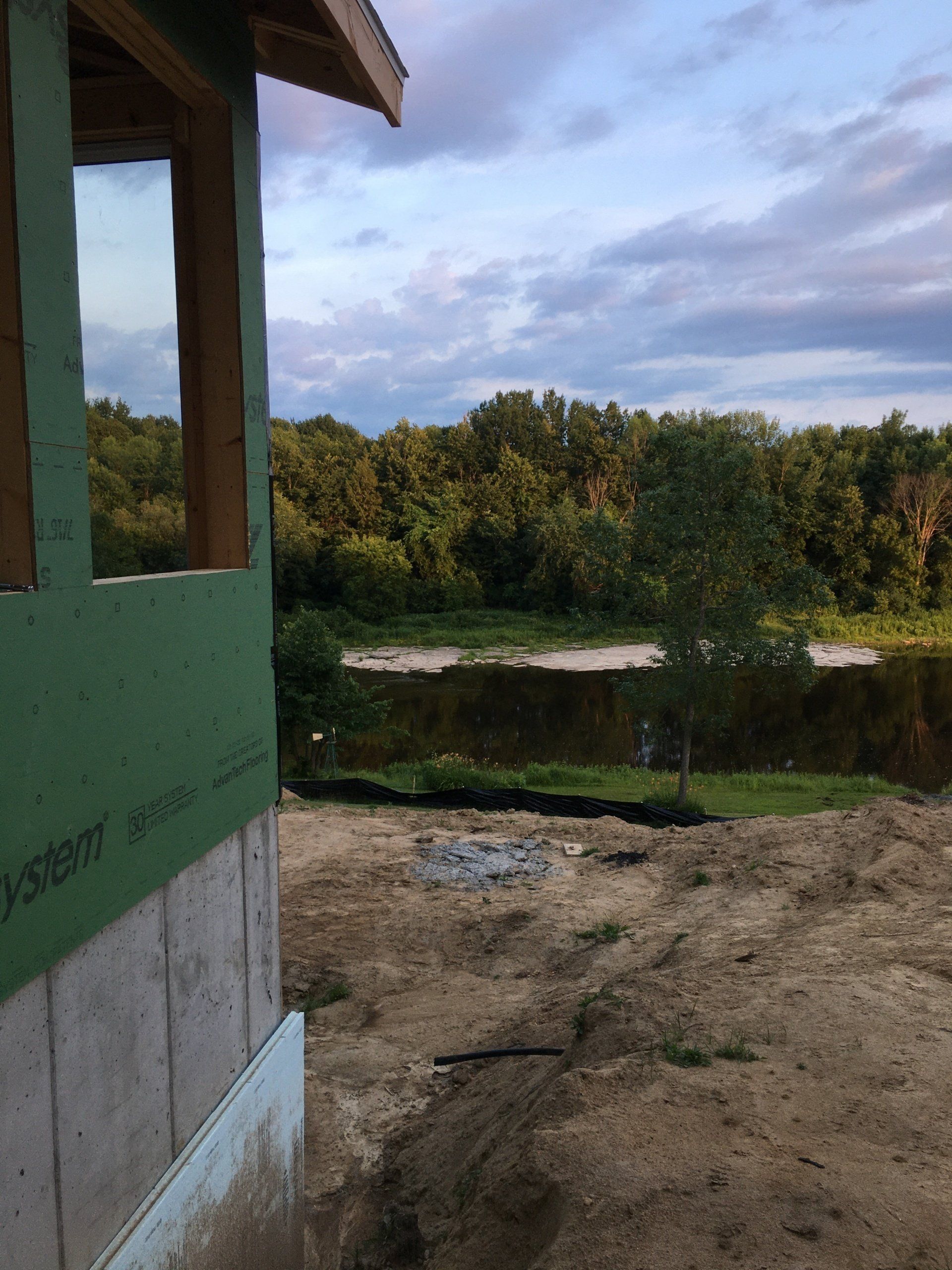 Partially built green house wall overlooking a dirt yard, pond, and trees under a cloudy sky
