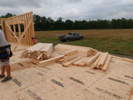 Wood-framed house under construction with lumber stacks on a foundation and a pickup truck in a field
