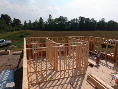 Wooden house framing under construction on a sunny lot, with lumber stacks and tools nearby
