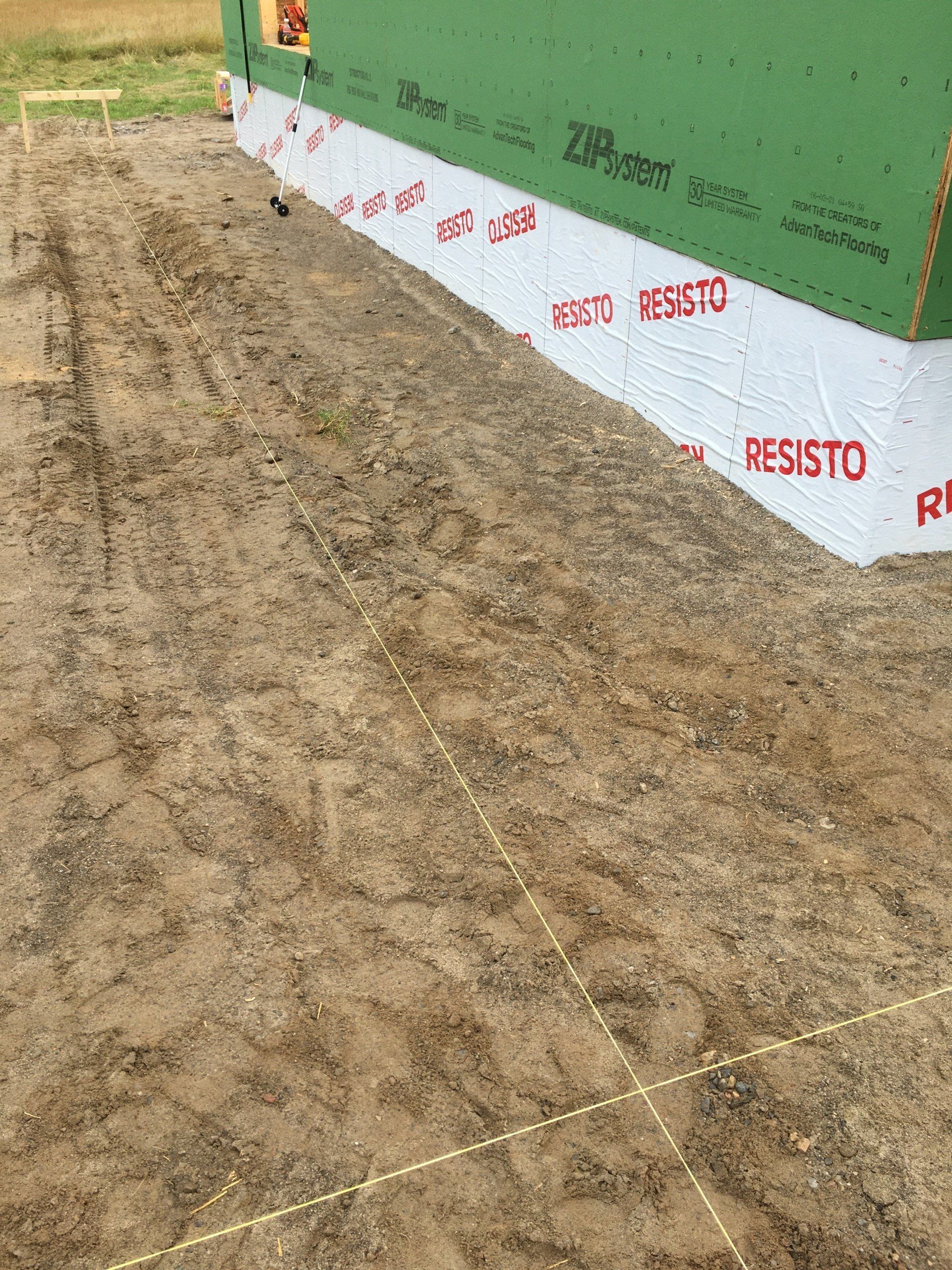 Bare dirt lot beside a green building wrapped with white-and-red RESISTO housewrap