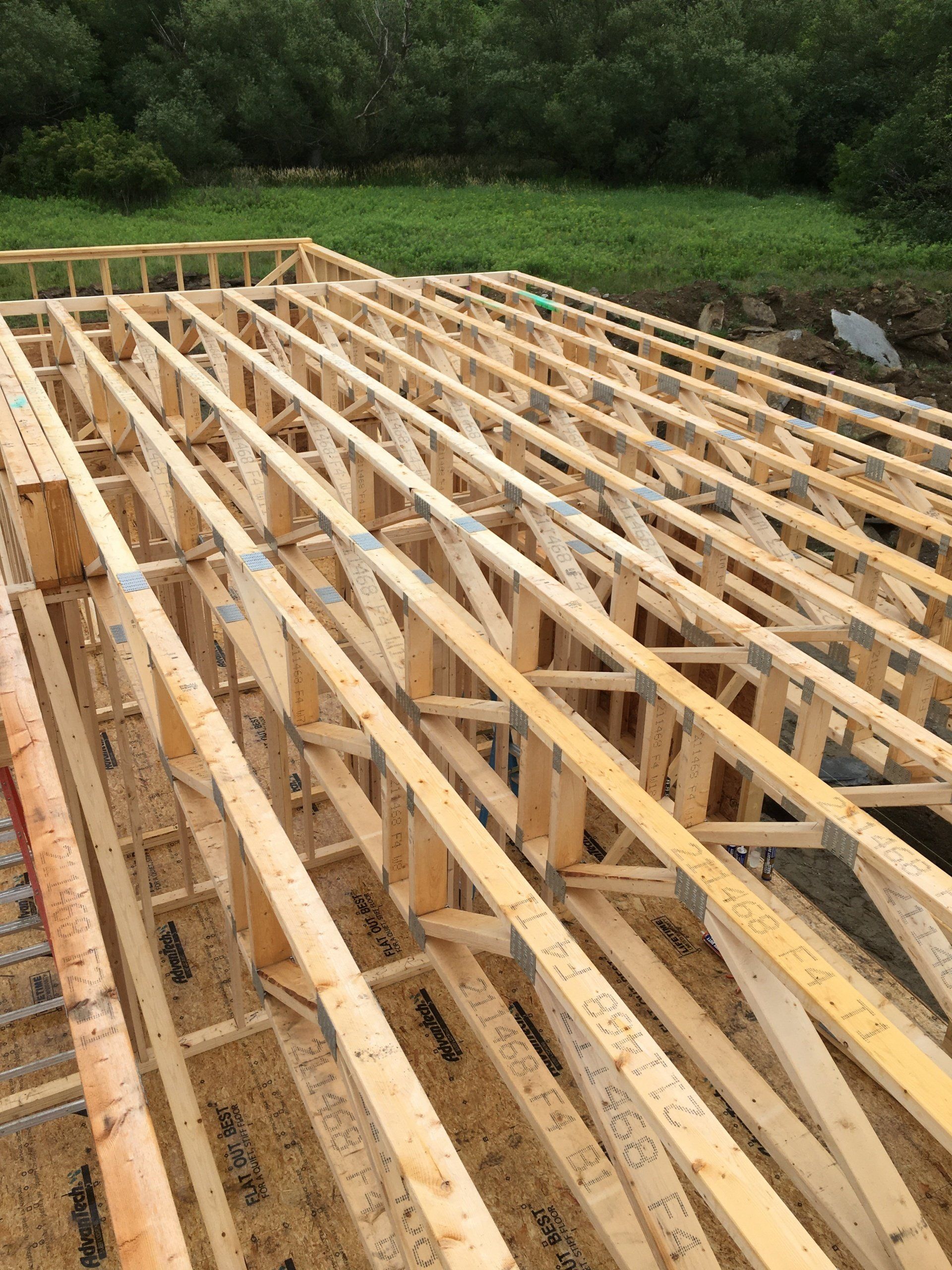 Wooden floor joists laid out in a construction site beside green trees and gravel.