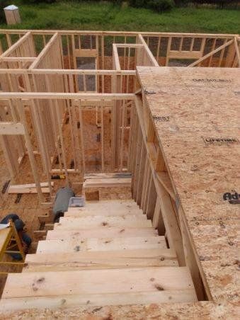 Wood-framed house under construction with plywood stairs and floor framing inside.