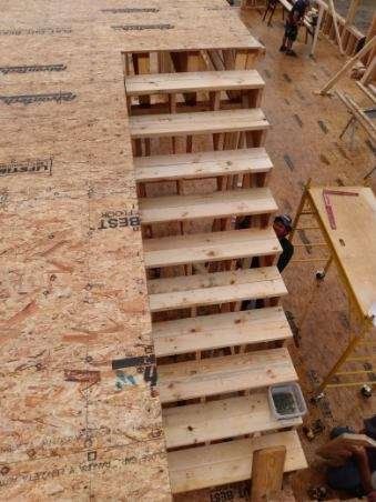 Wooden staircase under construction on a plywood floor, with tools and workers nearby.