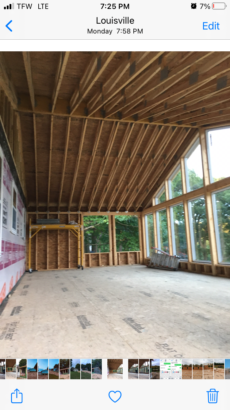 Empty unfinished room with exposed wood framing, sloped ceiling, and large windows overlooking trees.