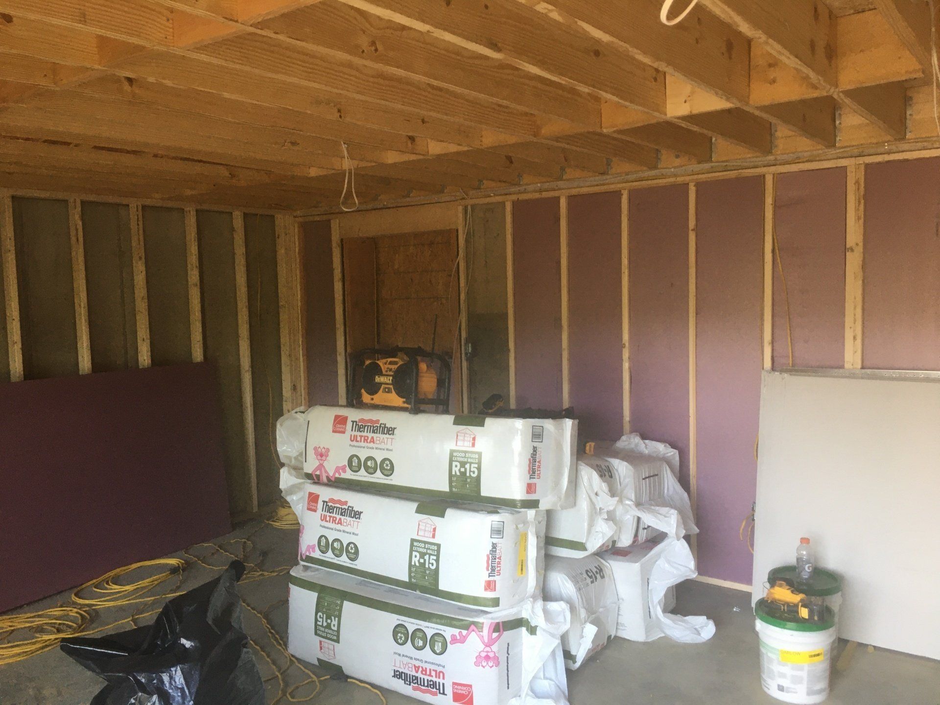 Unfinished room with exposed studs, insulation, stacked building materials, and a black bag on the floor.