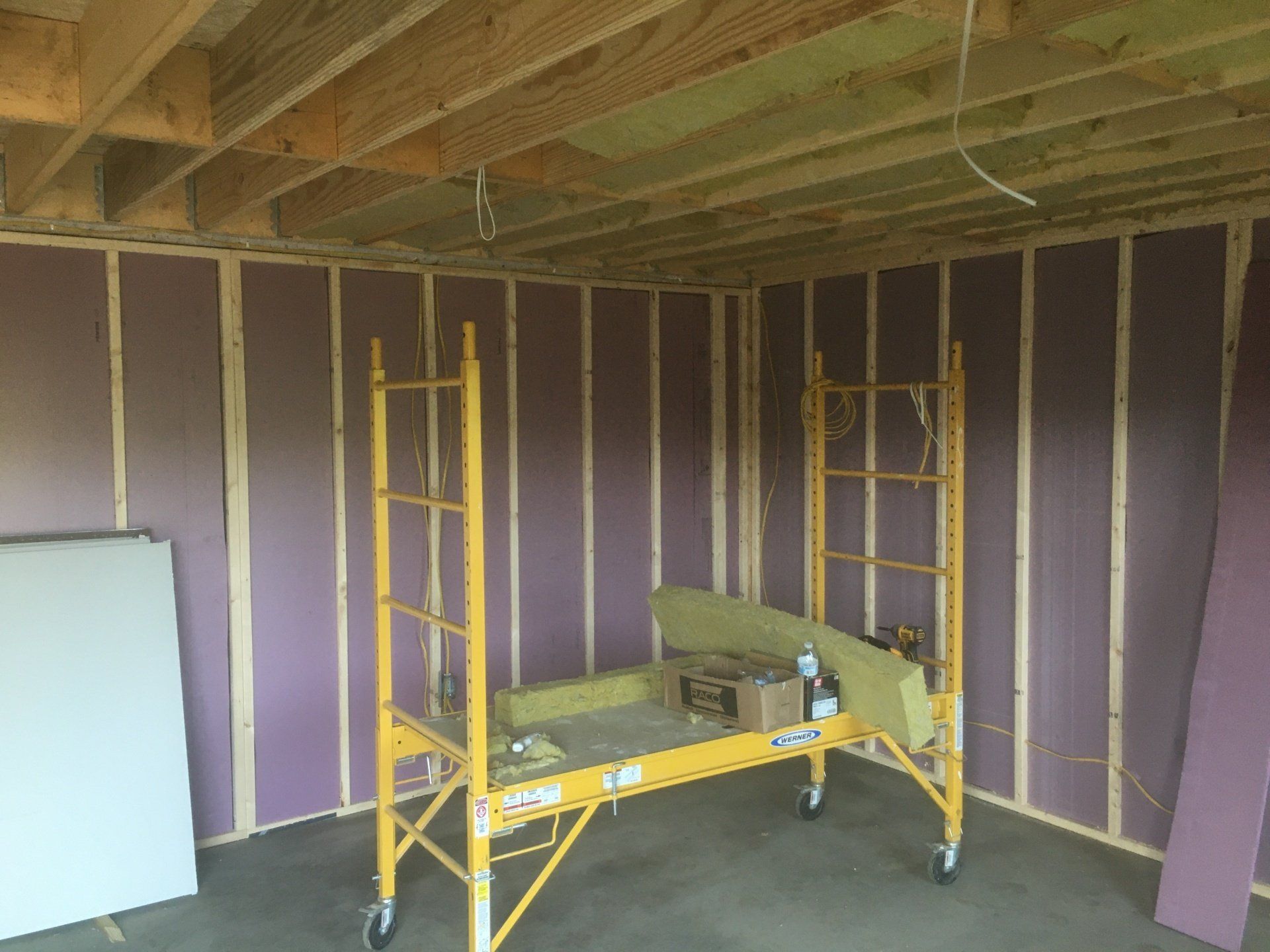 Basement under construction with purple insulation walls, exposed ceiling, and yellow scaffolding in the center