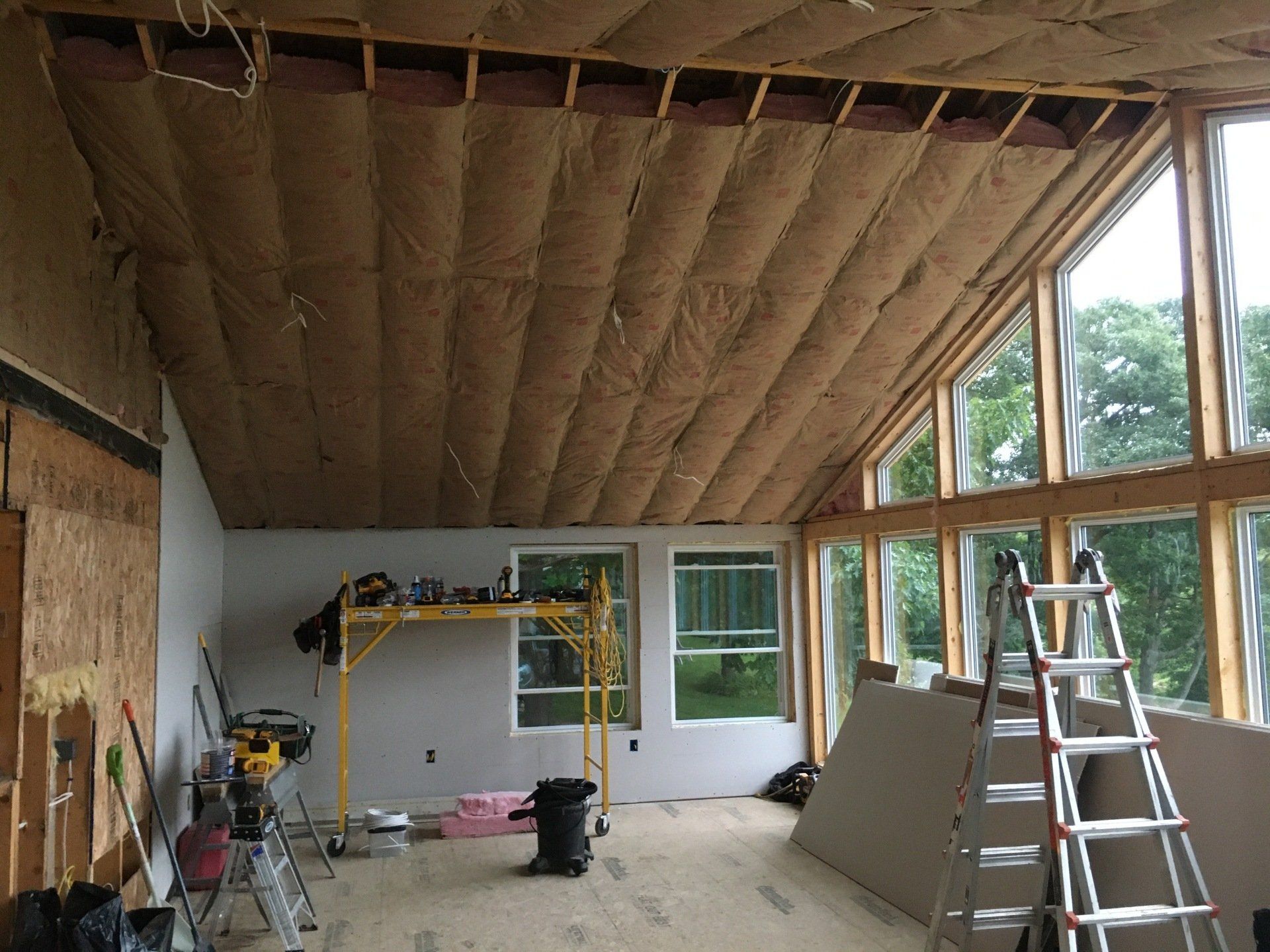 Interior room under construction with exposed insulation, windows, a ladder, and work tools.