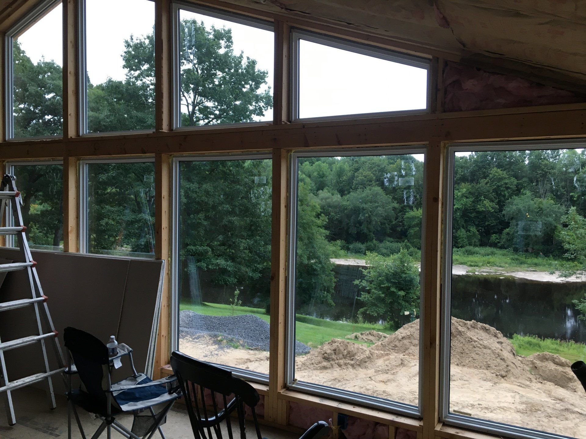 Partially finished room with large windows overlooking a green yard and pond, with a ladder and chairs inside