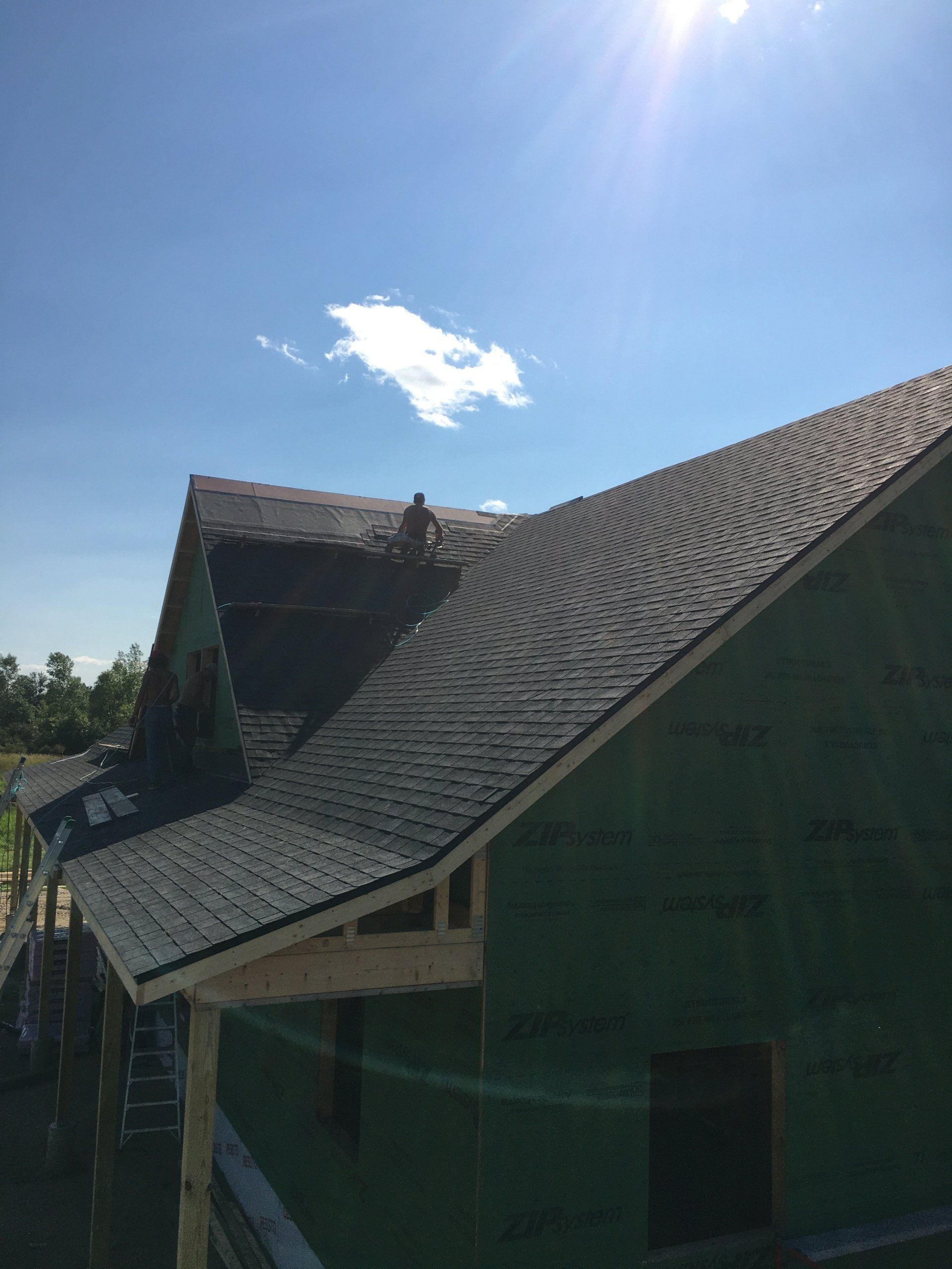 House roof under construction with dark shingles and green netting beneath a bright blue sky