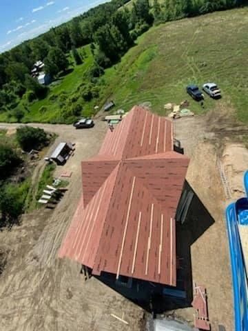 Aerial view of a red-roofed house on a dirt lot beside green trees and parked vehicles.