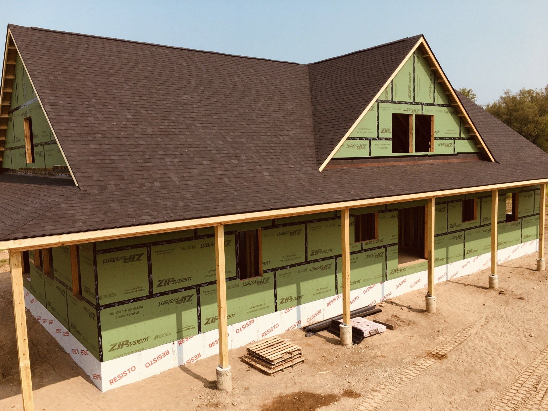 Partially built house with green sheathing, brown roof, and covered porch on a dirt lot