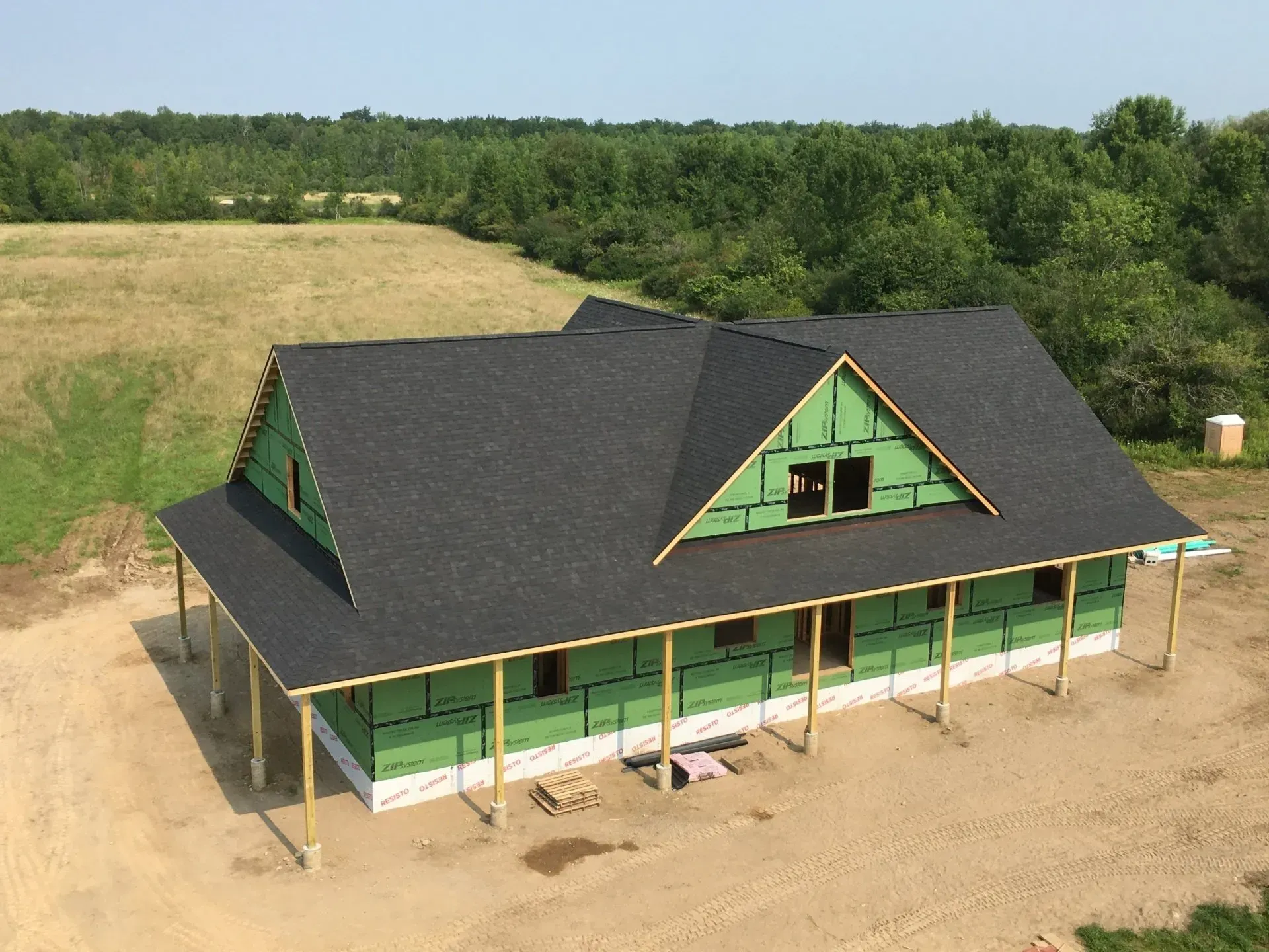 Green house under construction with black roof and wraparound porch in a rural field