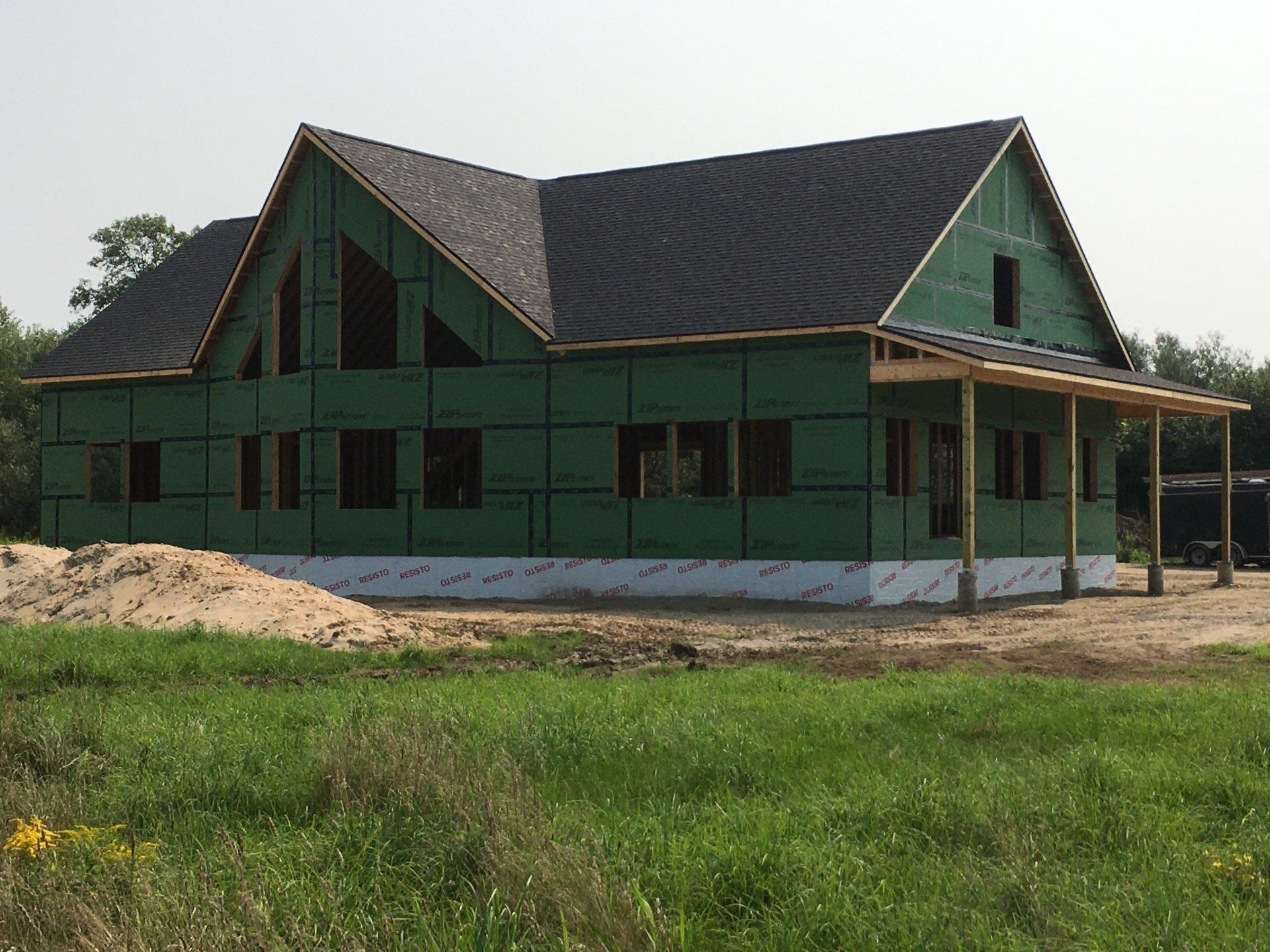 Green house under construction with dark roof, porch, and unfinished exterior in a grassy lot