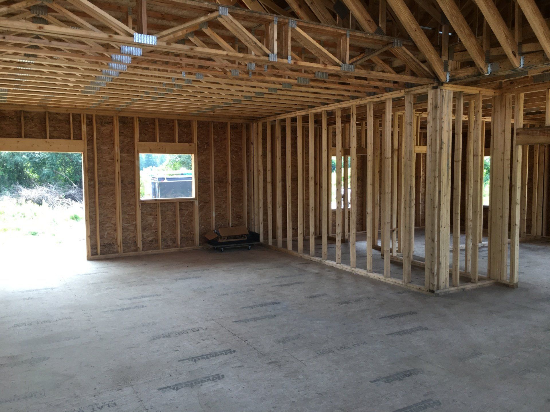 Unfinished wooden house interior with exposed framing, plywood roof trusses, and concrete floor.