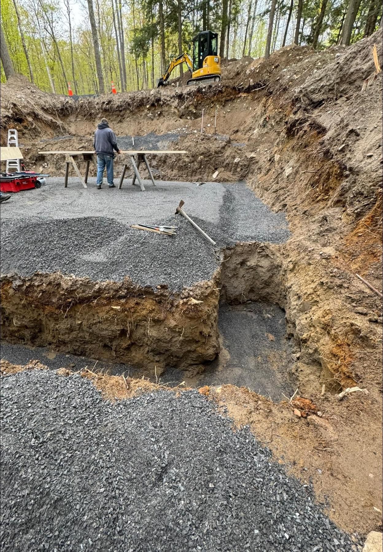Construction site with gravel foundation, workers, and excavator on a wooded slope