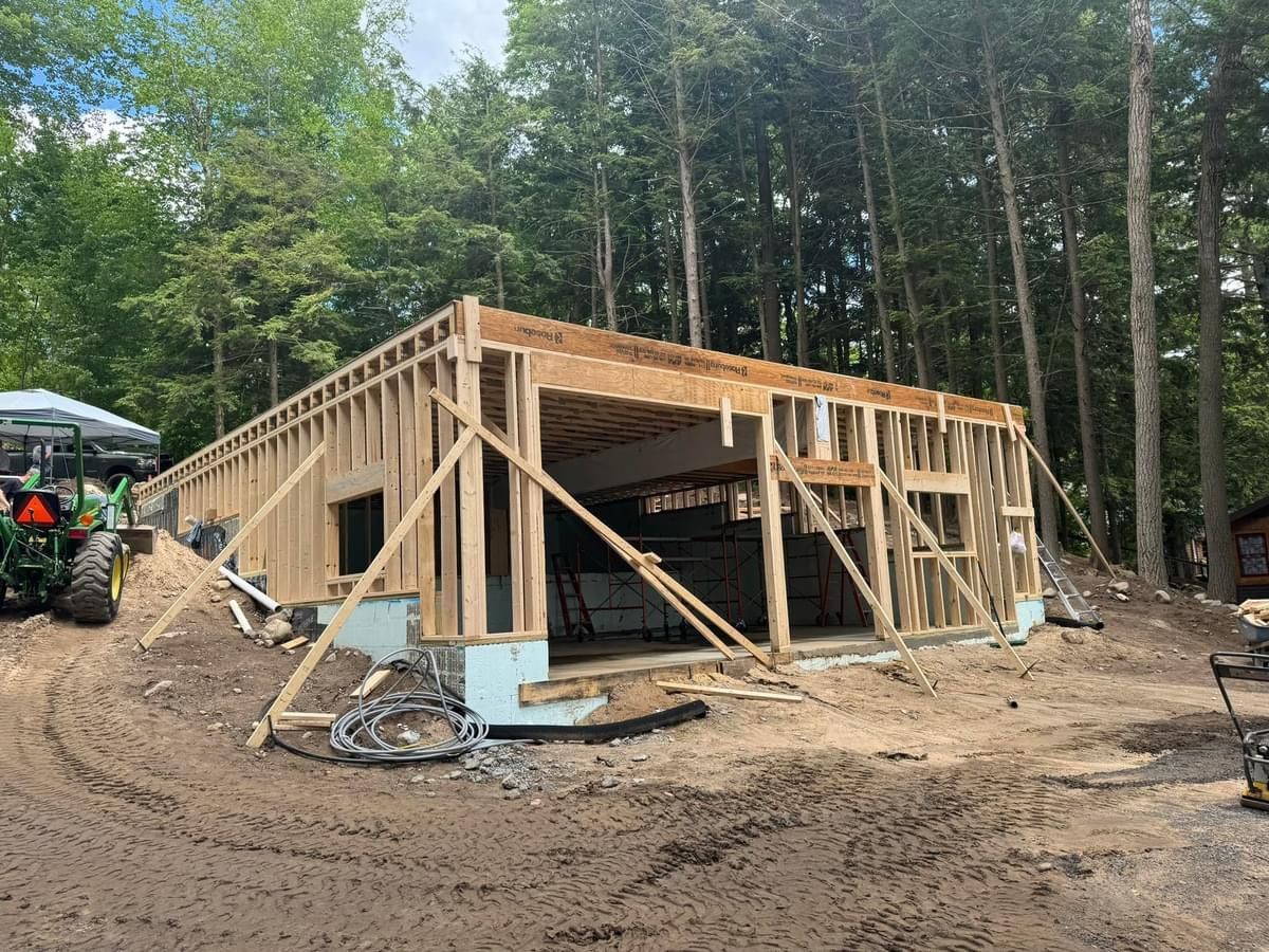 Wood-framed house under construction on a muddy site beside trees and a tractor