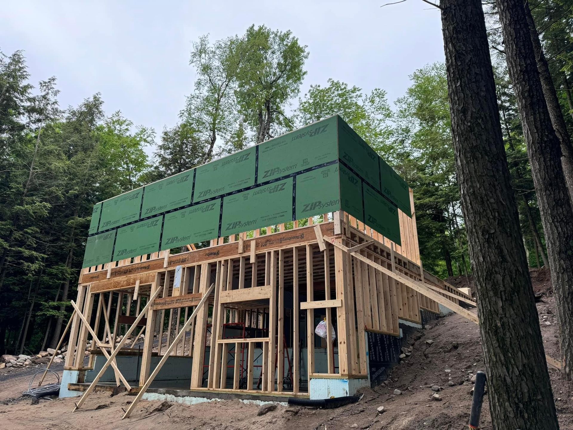 Wood-framed house under construction on a wooded hillside, with green protective sheathing on the upper level.