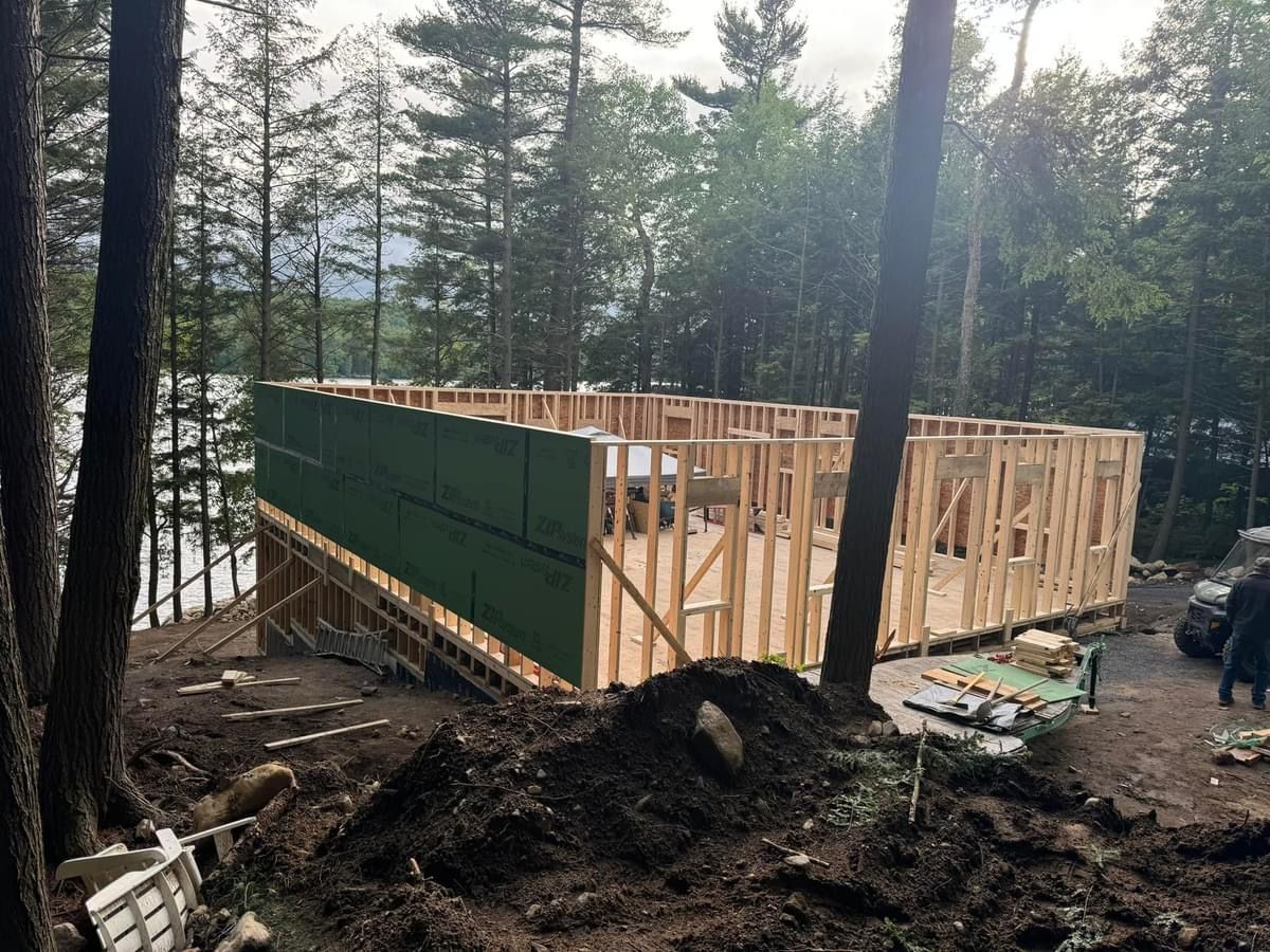 Wooden cabin frame under construction in a forest, with green sheathing and trees around it