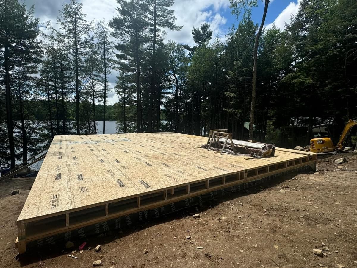 Partially built wooden deck platform beside a lake, surrounded by pine trees and construction materials