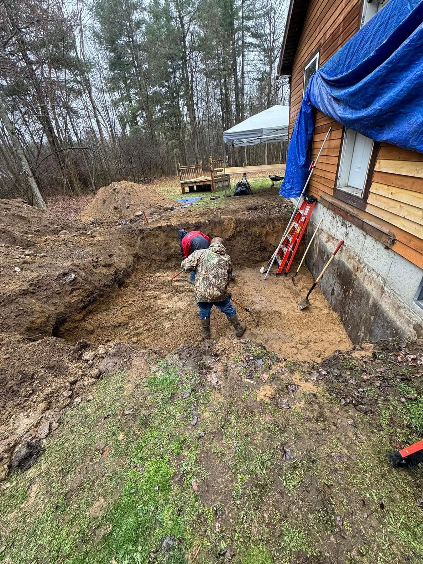 Worker digging beside house foundation near wooded yard, with blue tarp covering wall.