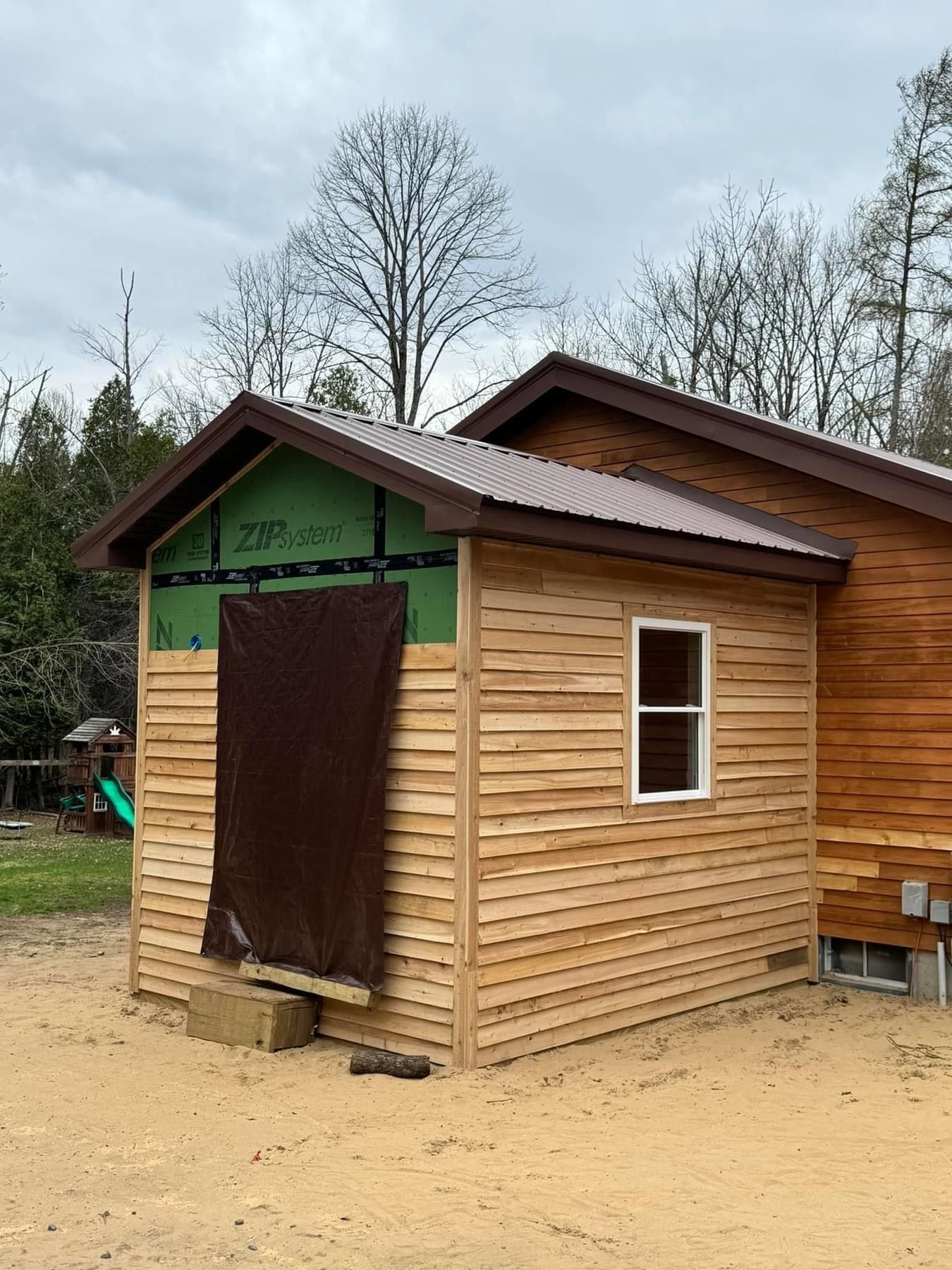 Small wooden cabin with a brown door covering the entrance, under a cloudy sky in a sandy yard.