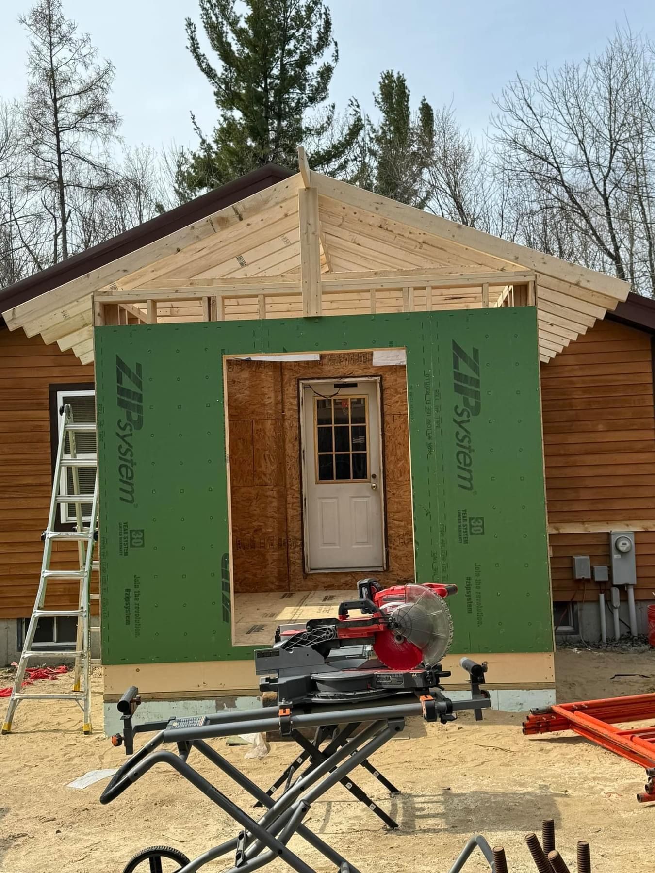 Small cabin entrance under construction with green sheathing, exposed roof truss, and a table saw in front.