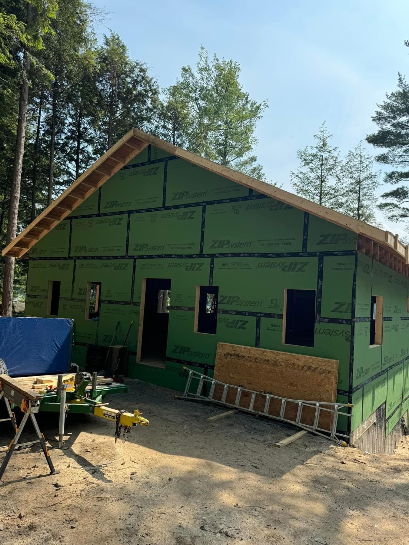 Partially built green house with exposed framing in a wooded area, surrounded by construction materials.