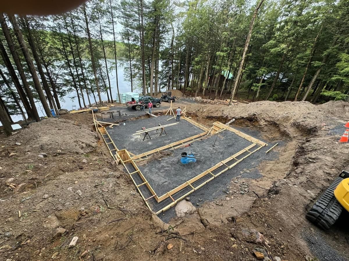 Construction site on a wooded lakeside slope with a foundation frame and gravel pad.