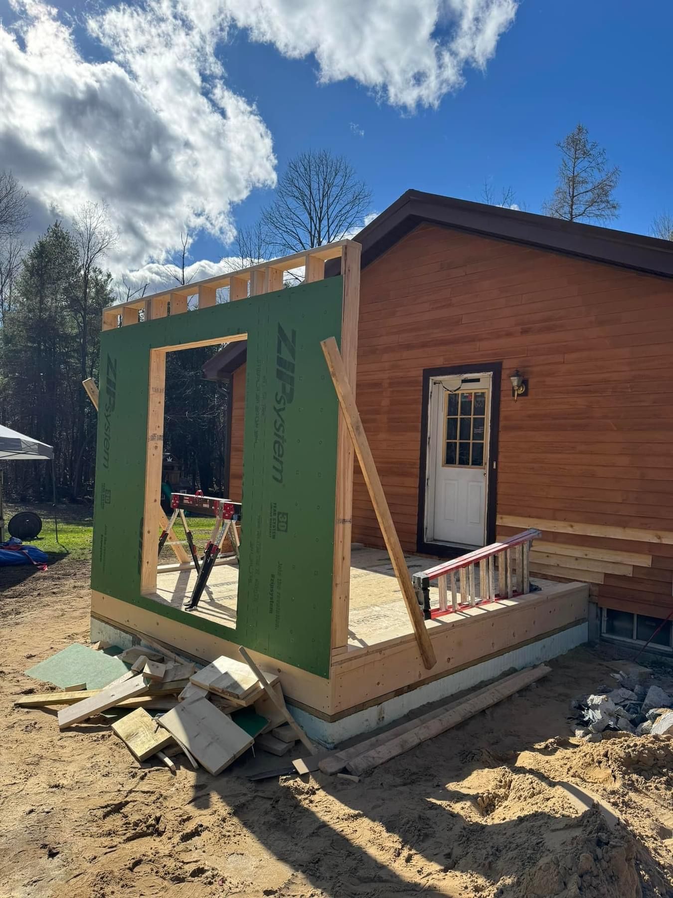 Partially built wooden house with green-framed opening and construction debris outside under a blue sky