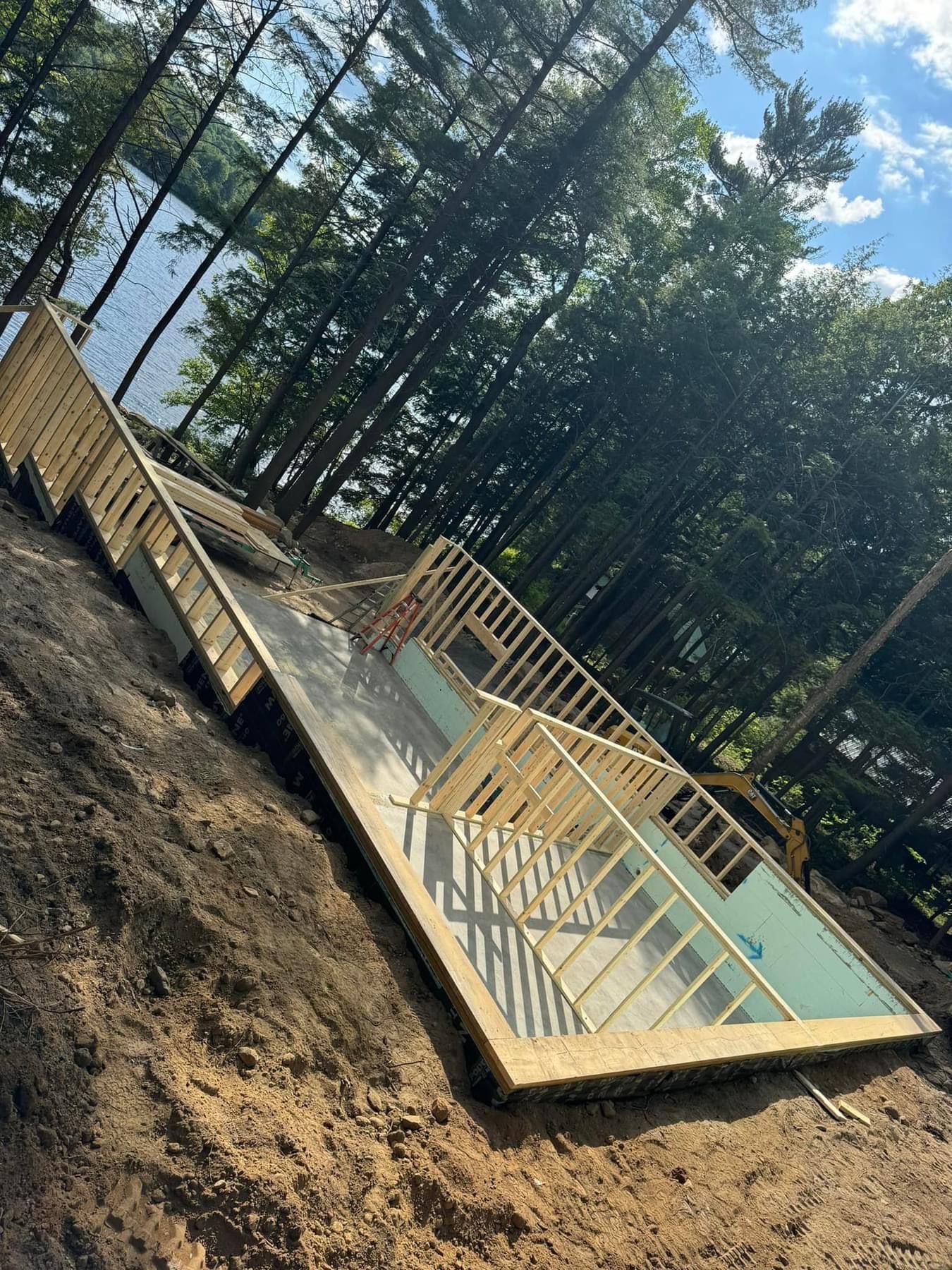 Wooden deck frame under construction in a wooded area, with yellow railings and a concrete slab.