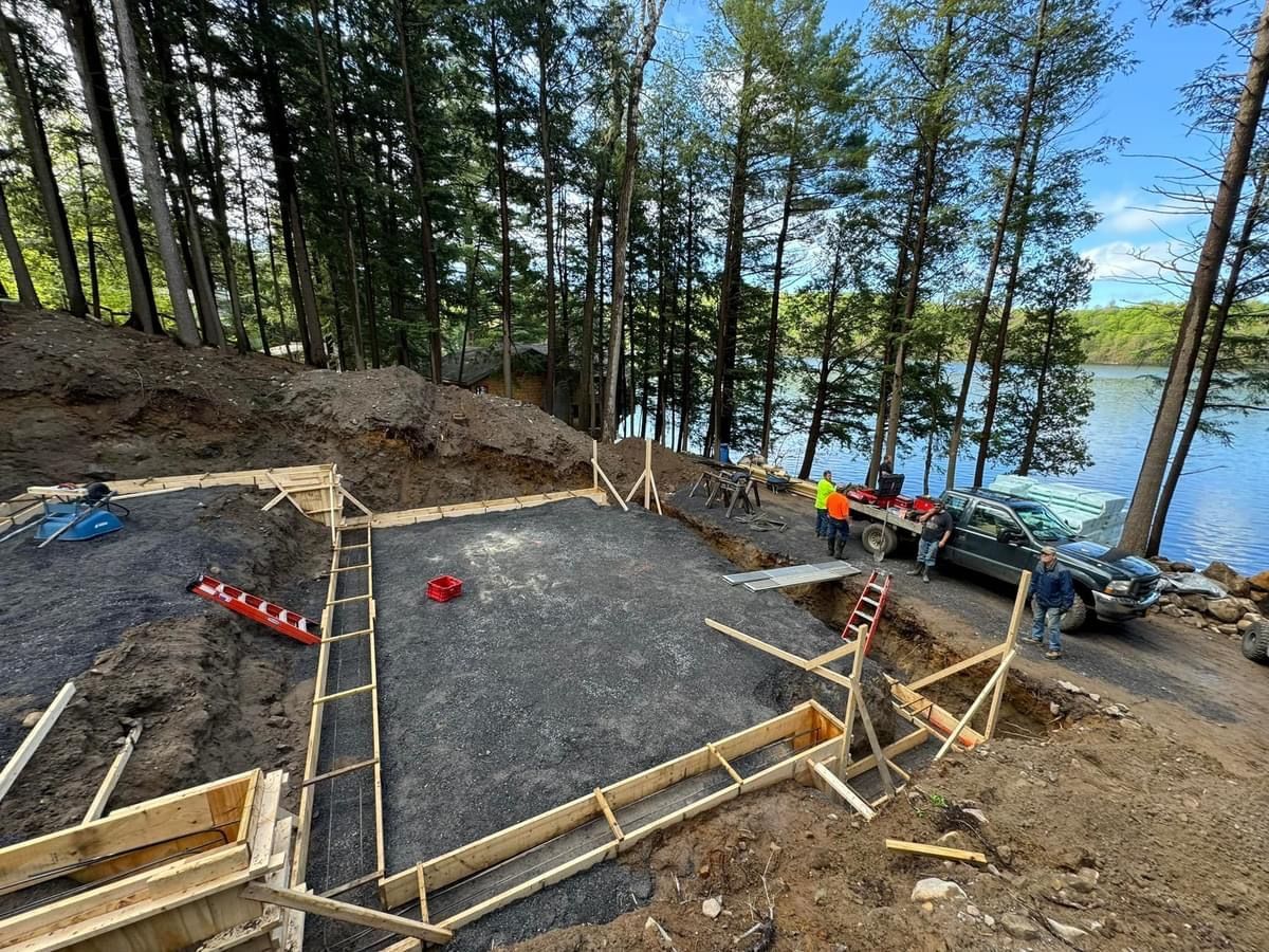 Construction site by a wooded lakeshore, with wooden forms and gravel foundation under a blue sky.