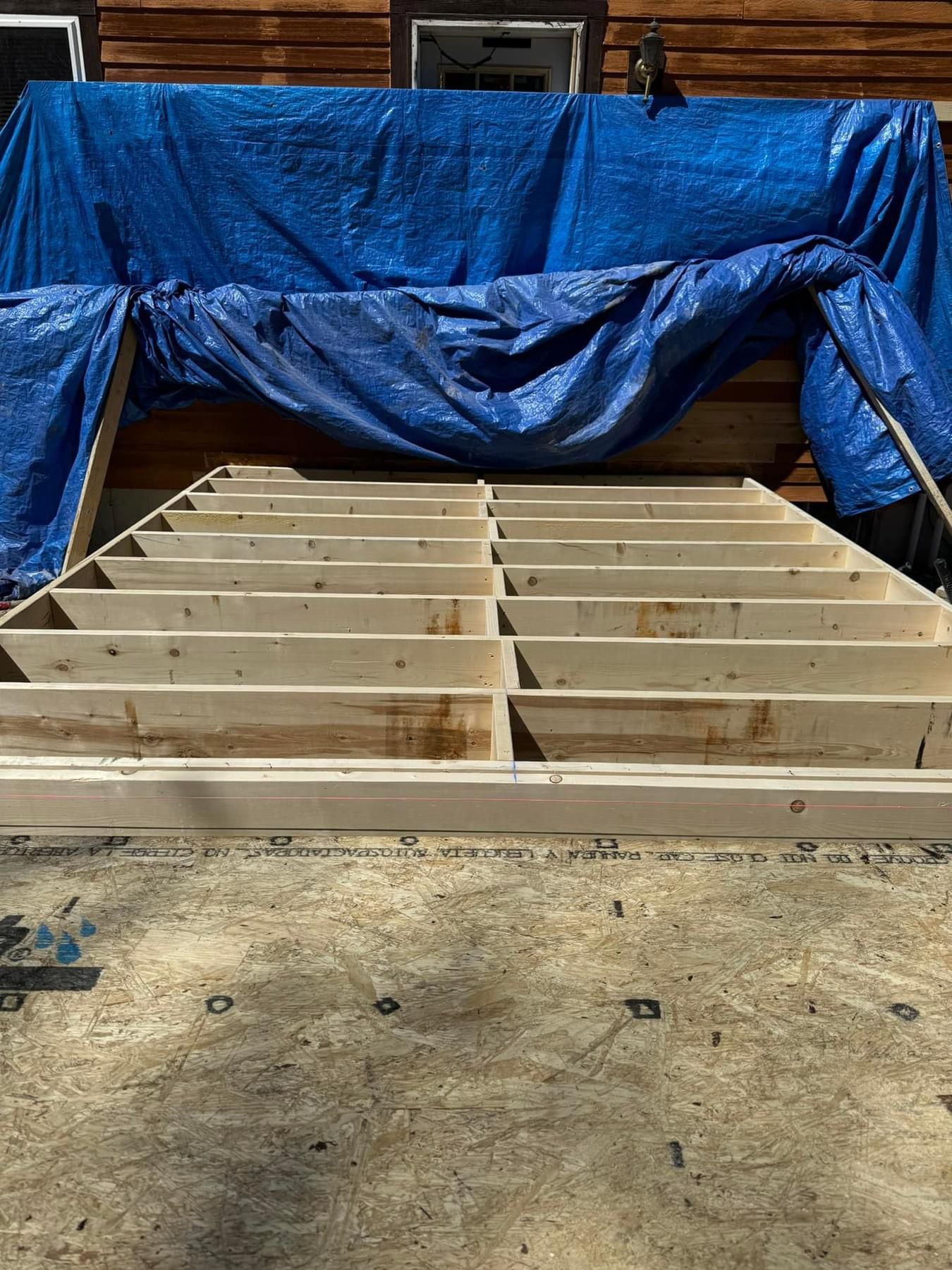 Wooden floor framing under construction, covered partly by a blue tarp in a backyard or worksite.