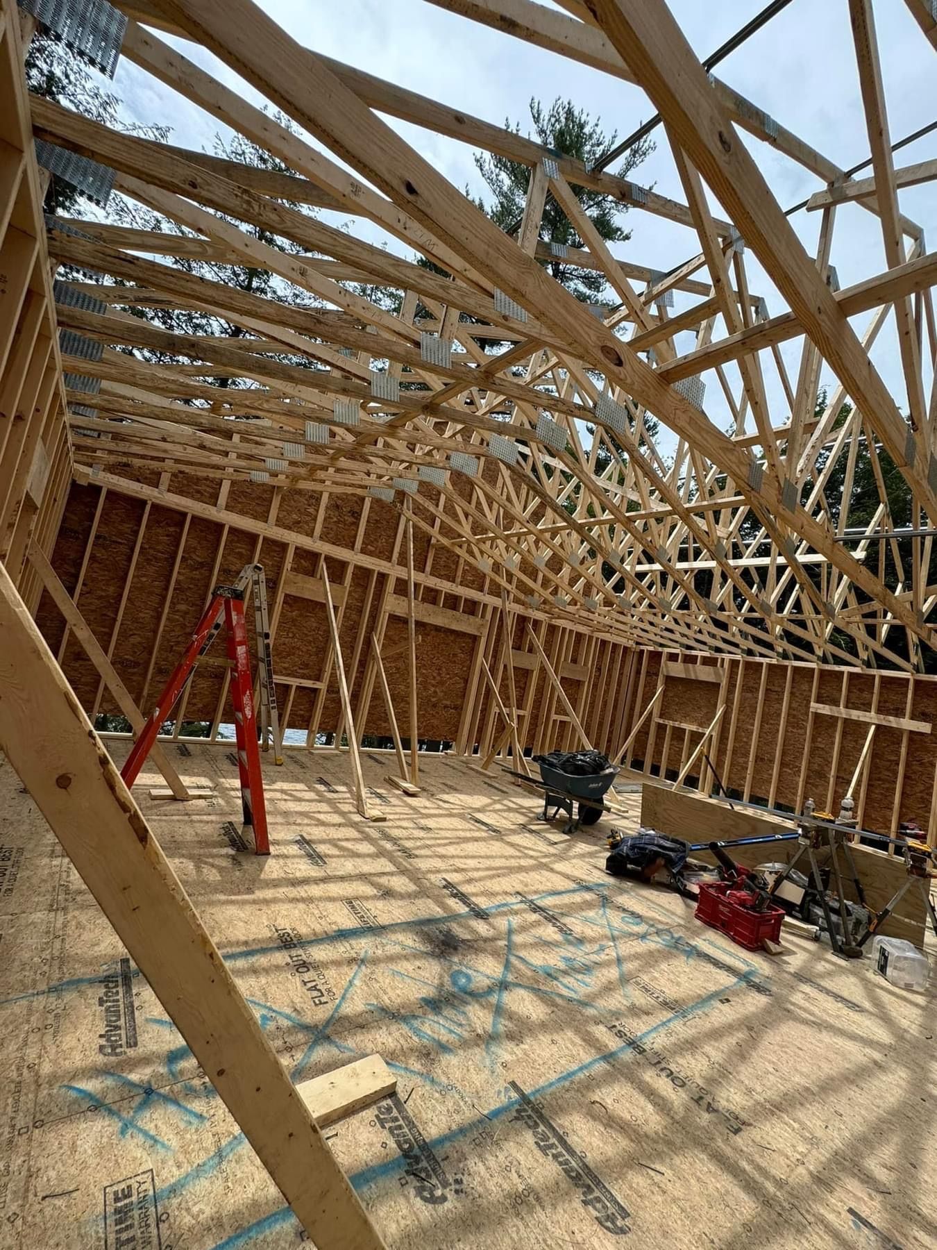 Wood-framed house under construction with roof trusses, red ladder, and tools on the floor