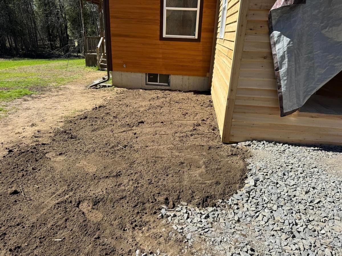 Fresh dirt beside a wooden shed and house, bordered by white gravel in a yard.