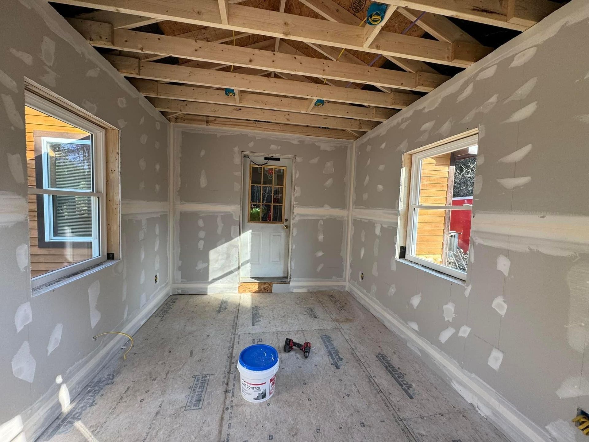 Partially finished room with drywall, exposed ceiling beams, two windows, and a paint bucket on the floor