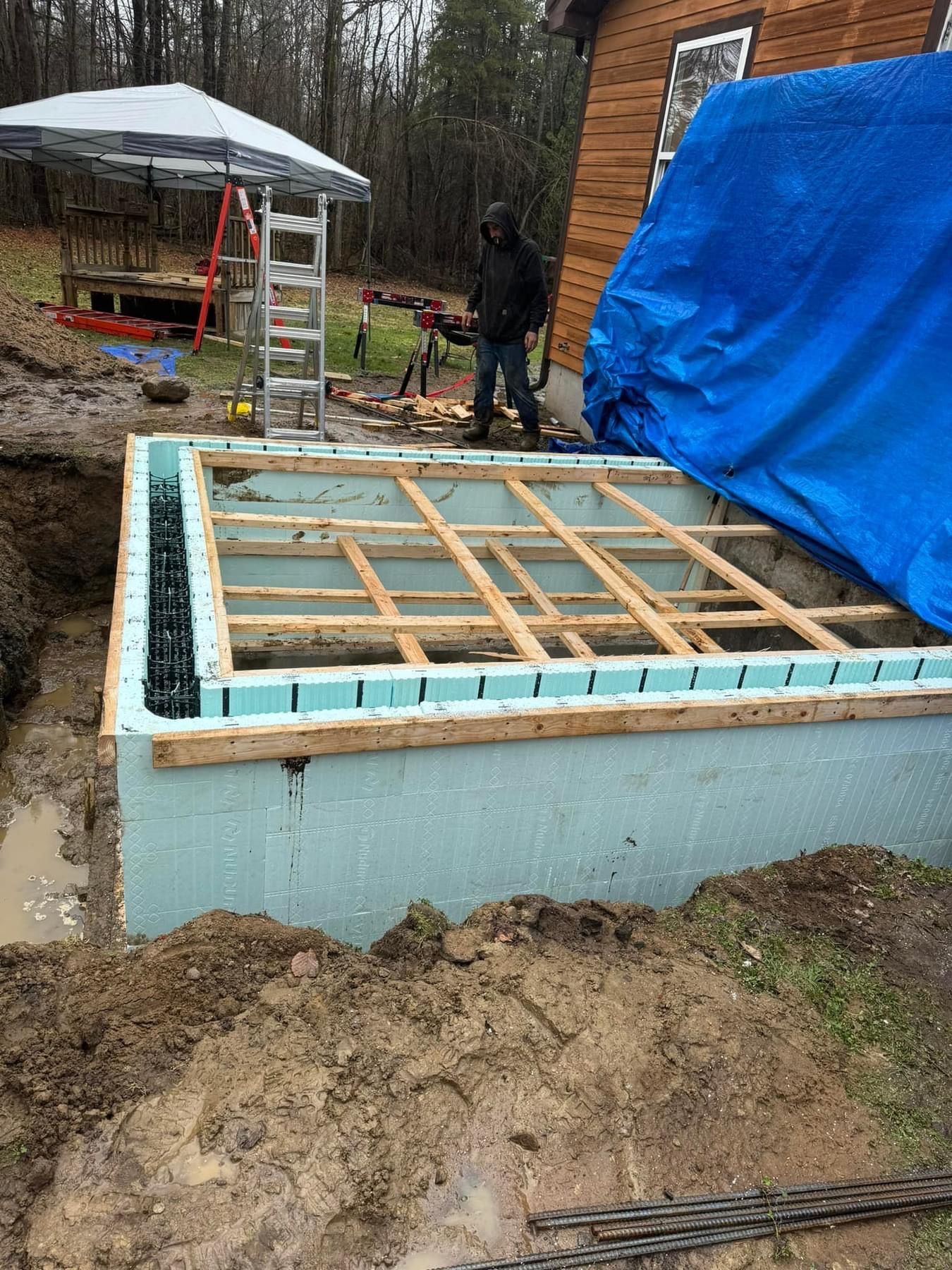 Wooden foundation framing under construction beside a blue tarp-covered structure in a muddy yard