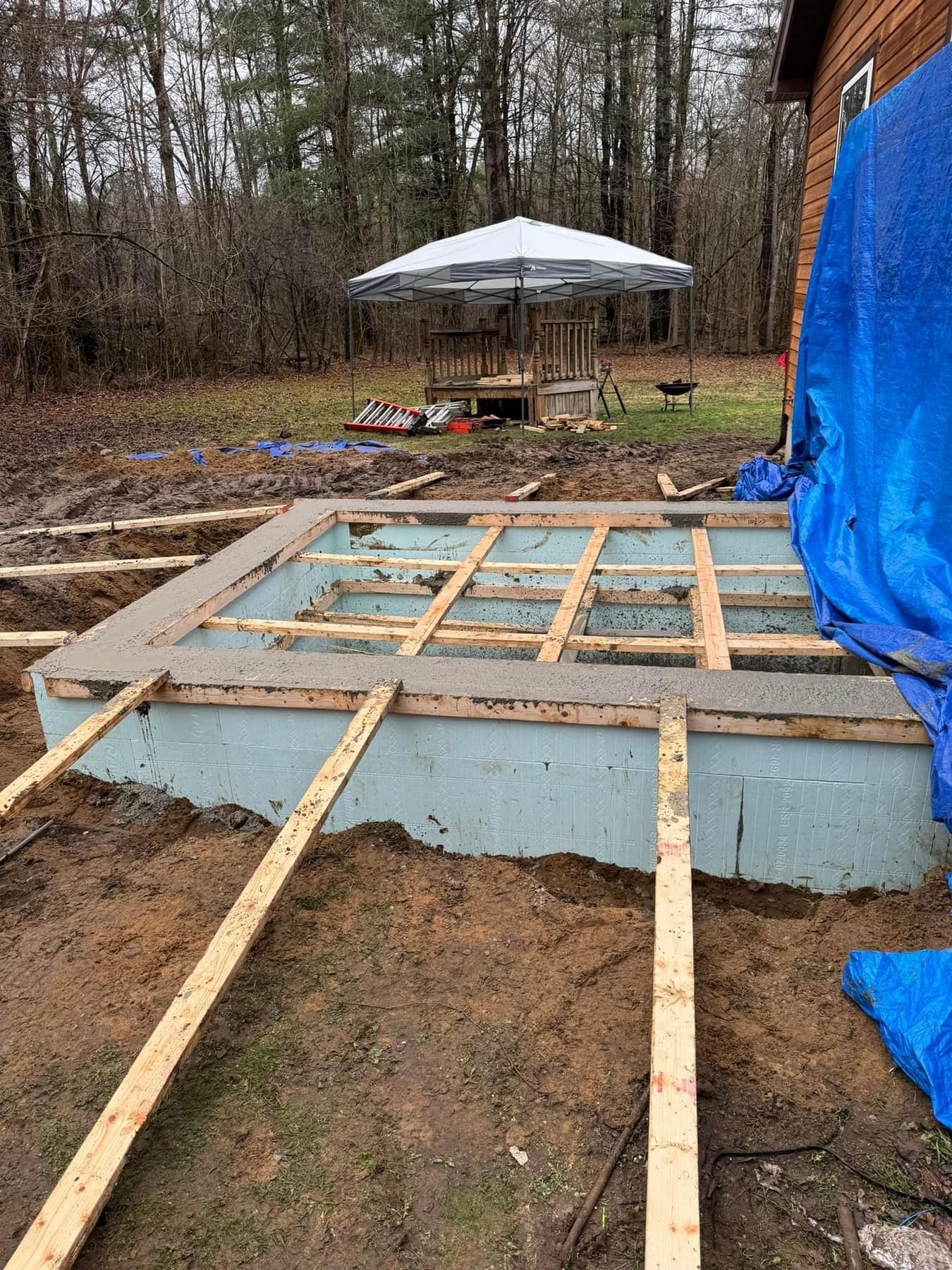 Partially built wooden deck frame on a raised concrete base in a muddy yard, with a tarp and patio umbrella nearby
