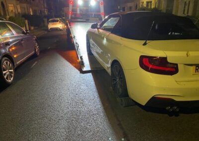 A white car is being towed down a street by a tow truck.