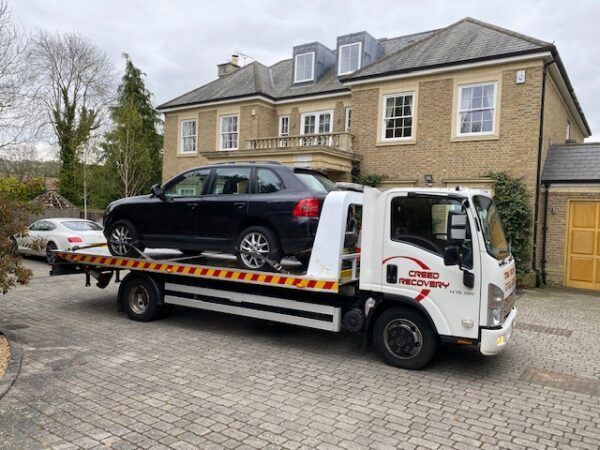 A tow truck with a car on the back is parked in front of a large house.