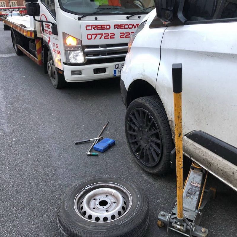 A white van with a flat tire being assisted by a recovery truck, tools laid out.