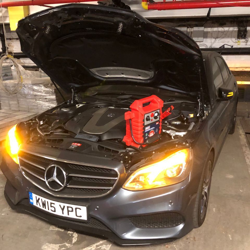 A grey Mercedes-Benz with its hood open and a jump starter on the engine, yellow hazard lights on.