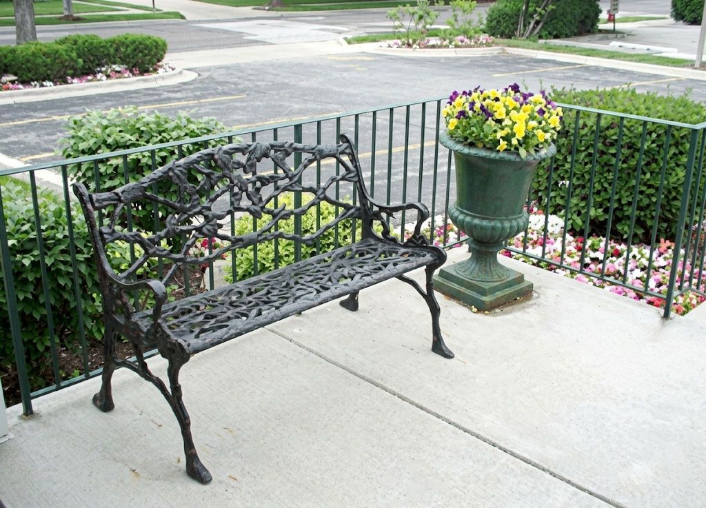 Black metal bench beside green planter with yellow flowers on a front porch.