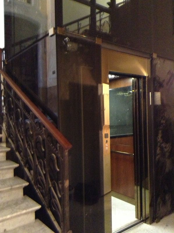 Vano scale poco illuminato accanto a un ascensore aperto con interni rivestiti in legno e cancello in metallo.