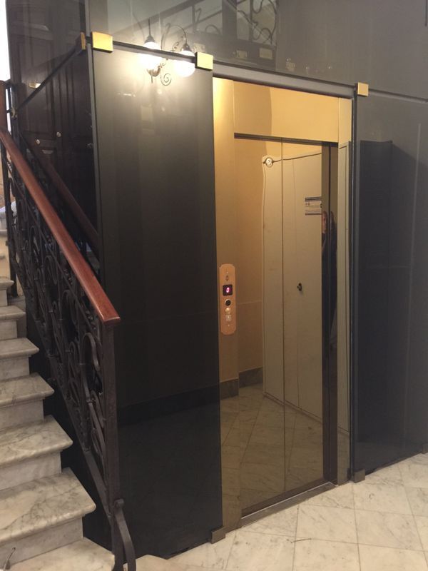 Ascensore in vetro accanto a una scala nella hall di un edificio, con pareti nere e interni illuminati.